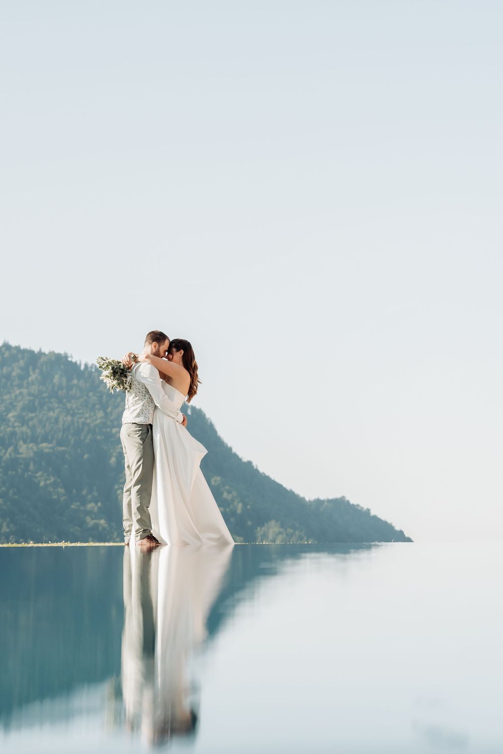 A romantic wedding couple embraces against a misty mountain lake backdrop in soft lighting.