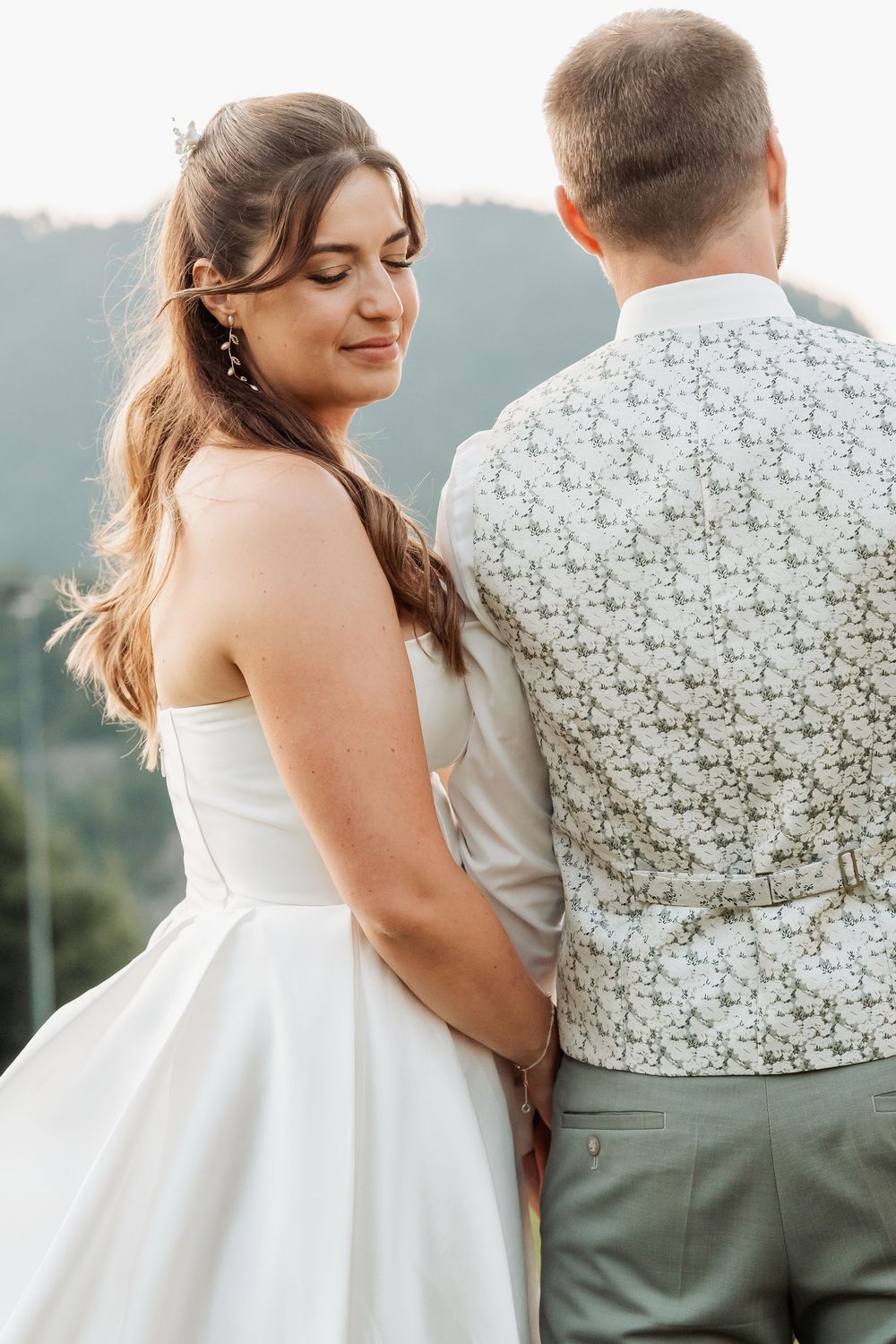 A bride in white gown and groom in patterned vest share an intimate moment against mountain scenery.