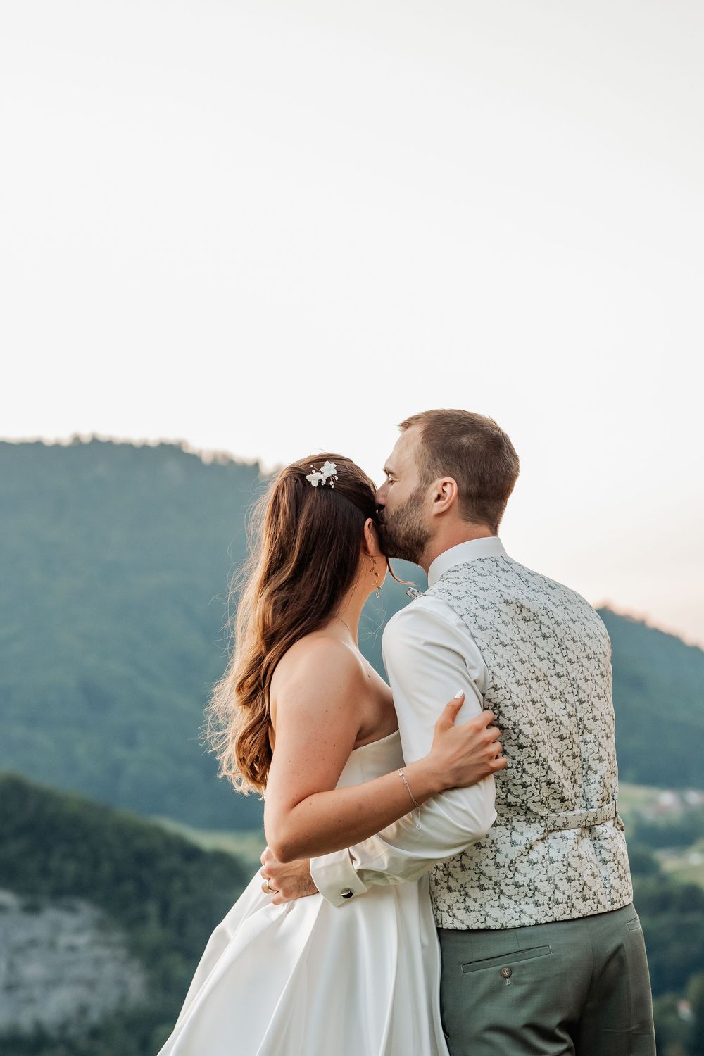 Newlyweds share a tender kiss while sitting together with mountain views in the background.