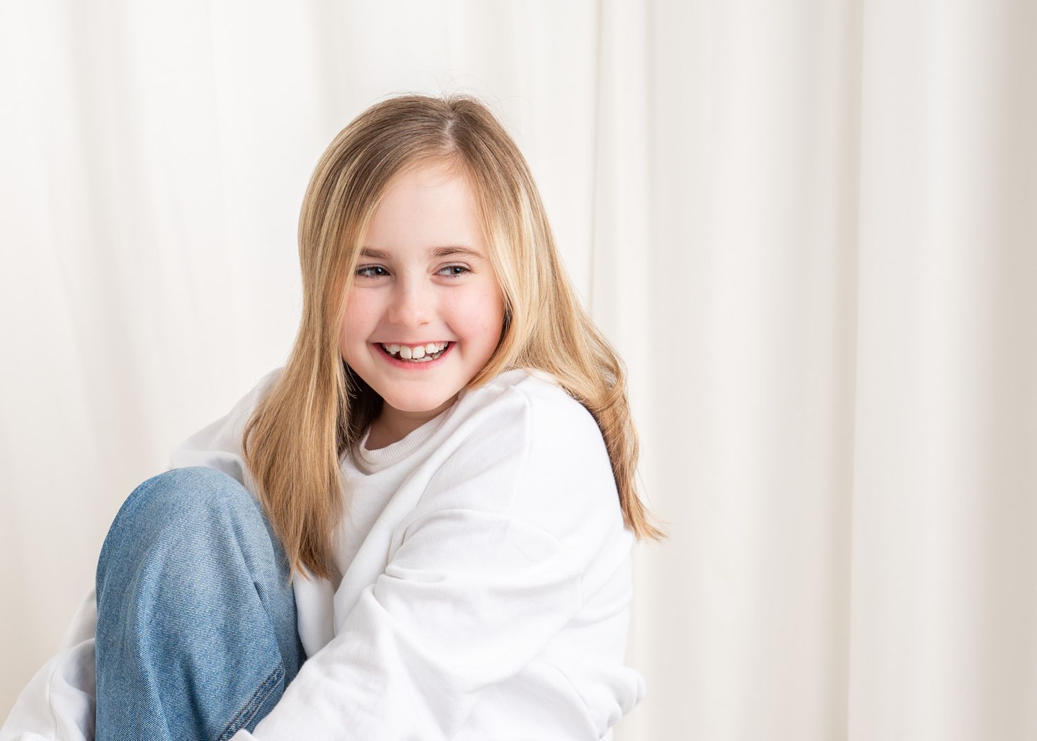 A person wearing a white sweater sits on a couch and smiles warmly while looking at the camera. TV headshots.