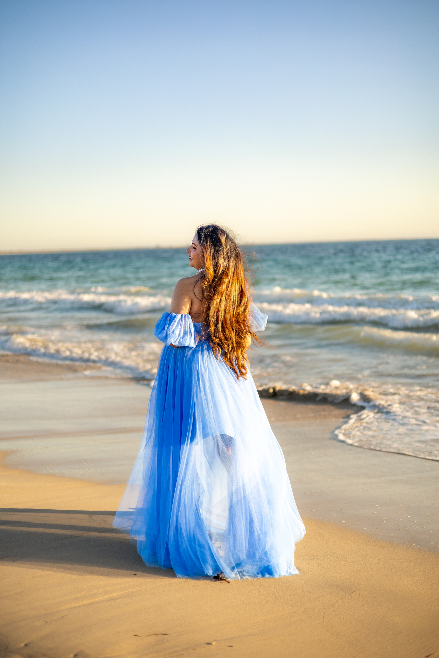 Two people in flowing blue and white dresses walk together along a sandy beach at sunset by the ocean waves.