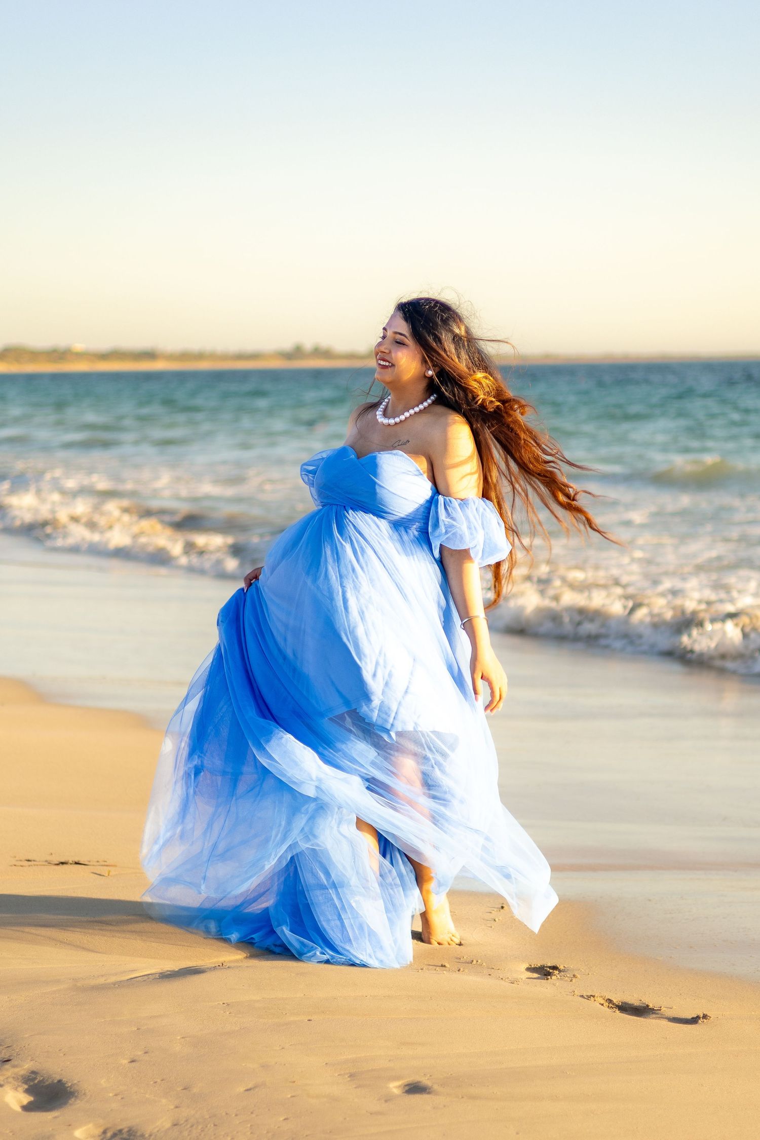 A figure in a flowing blue and white dress crouches on a sandy beach at sunset with waves in the background.