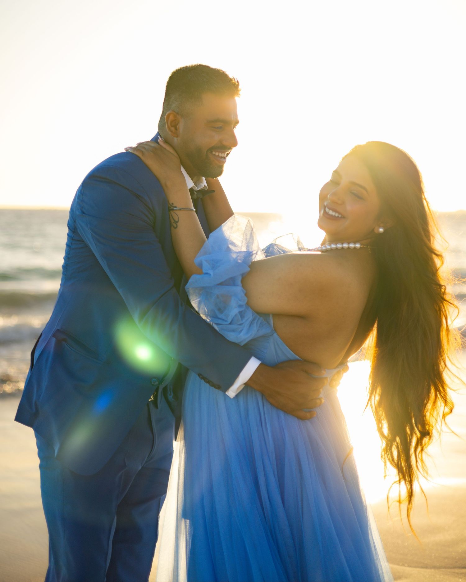 A romantic couple in blue attire embraces on a beach at sunset with sunflare creating a dreamy atmosphere.