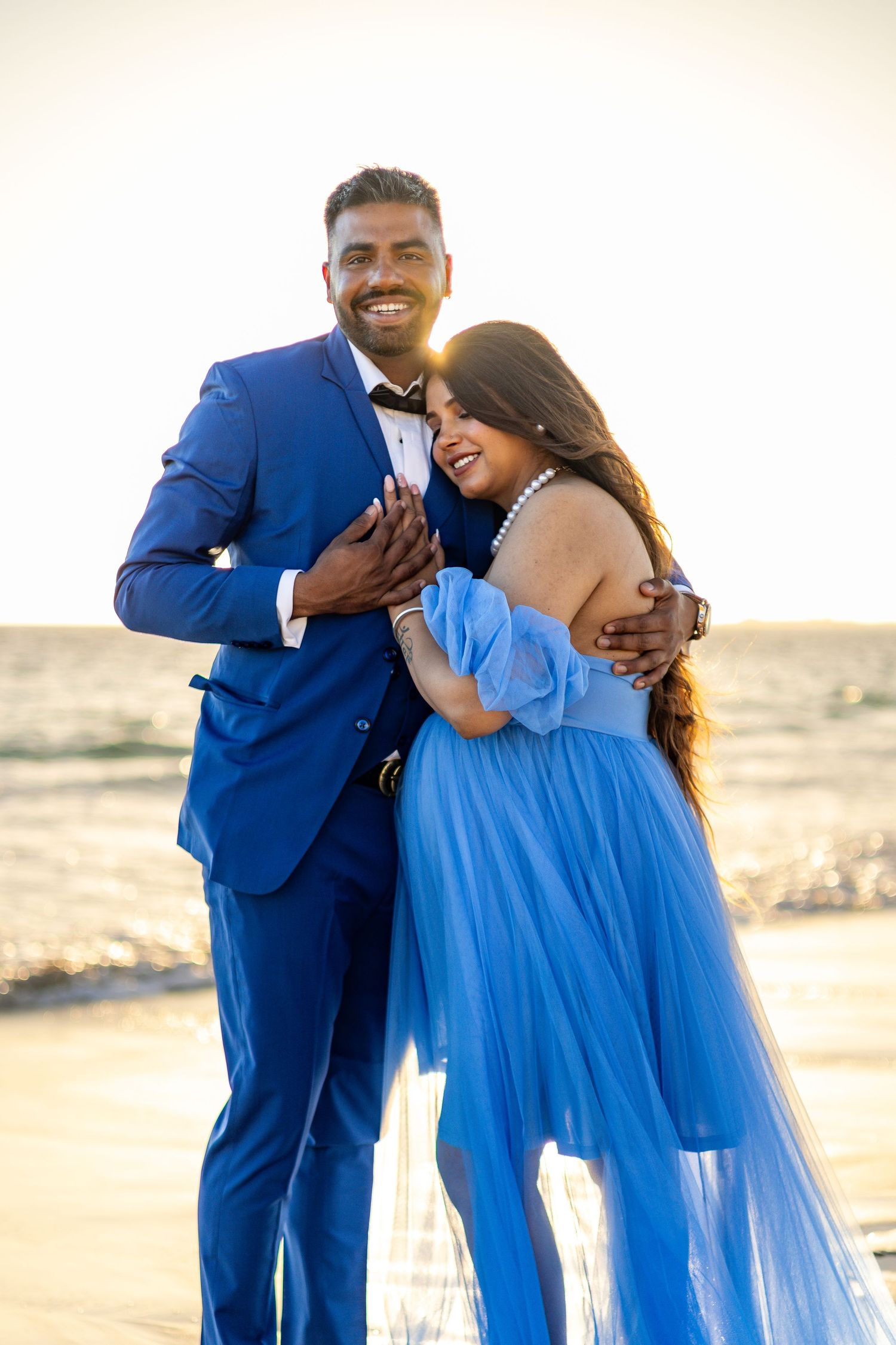 A couple embraces during a sunset photoshoot, with one wearing a royal blue suit and the other in a flowing light blue dress.
