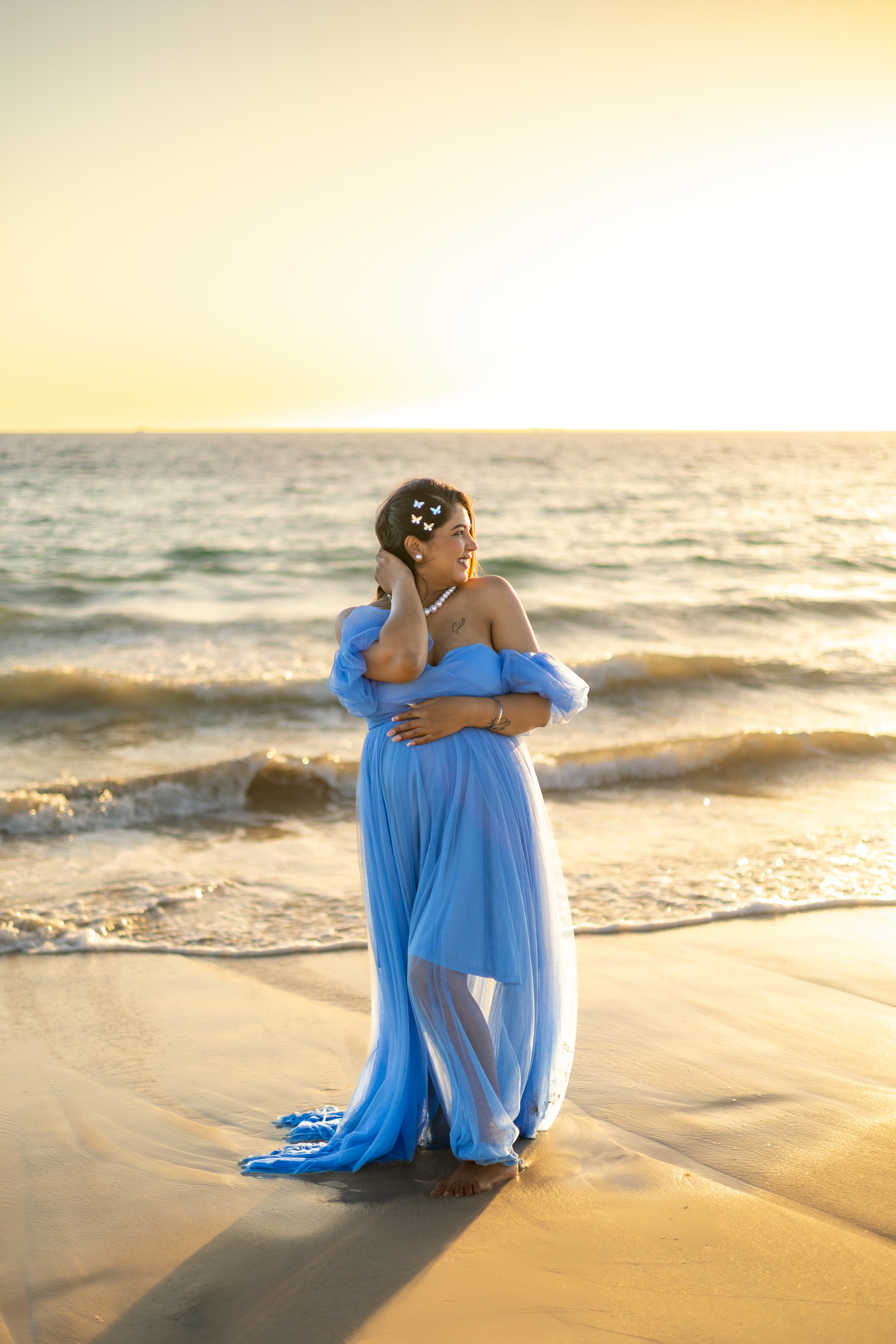 Person in flowing blue dress sits peacefully on golden beach at sunset as waves roll in along shoreline.