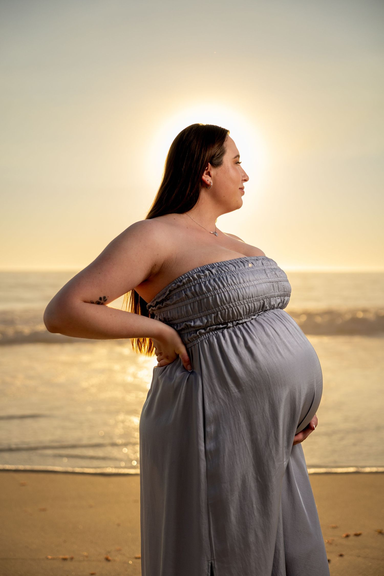 Expectant mother in gray dress poses on sandy beach during golden hour photoshoot.