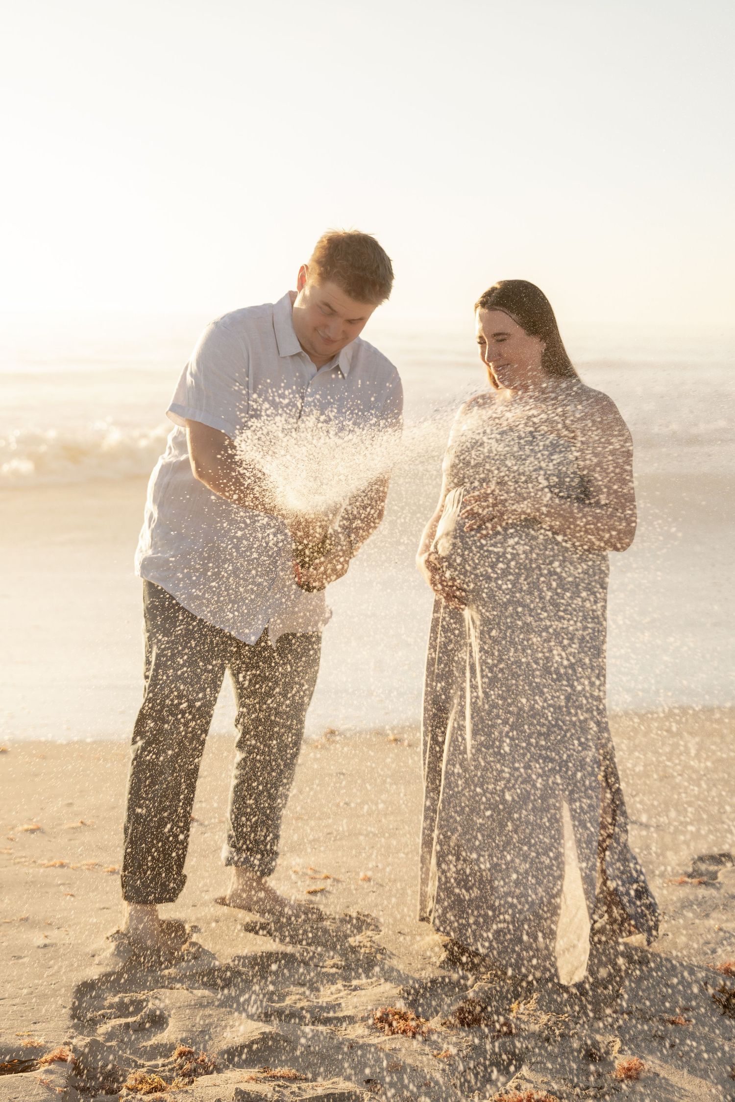 Couple playfully throwing sand at each other on a sunlit beach during golden hour.