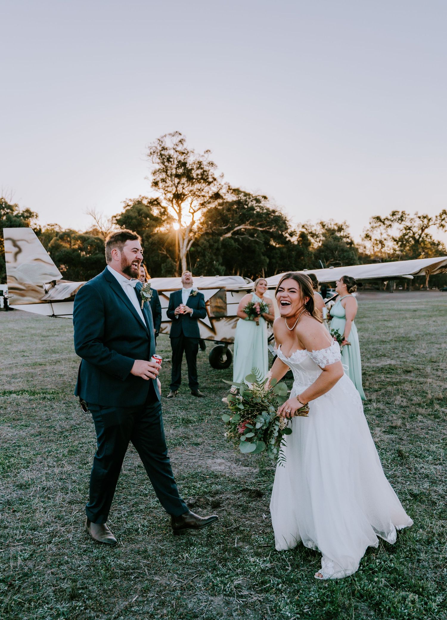 Wedding couple dances playfully outdoors in a grassy field at sunset.