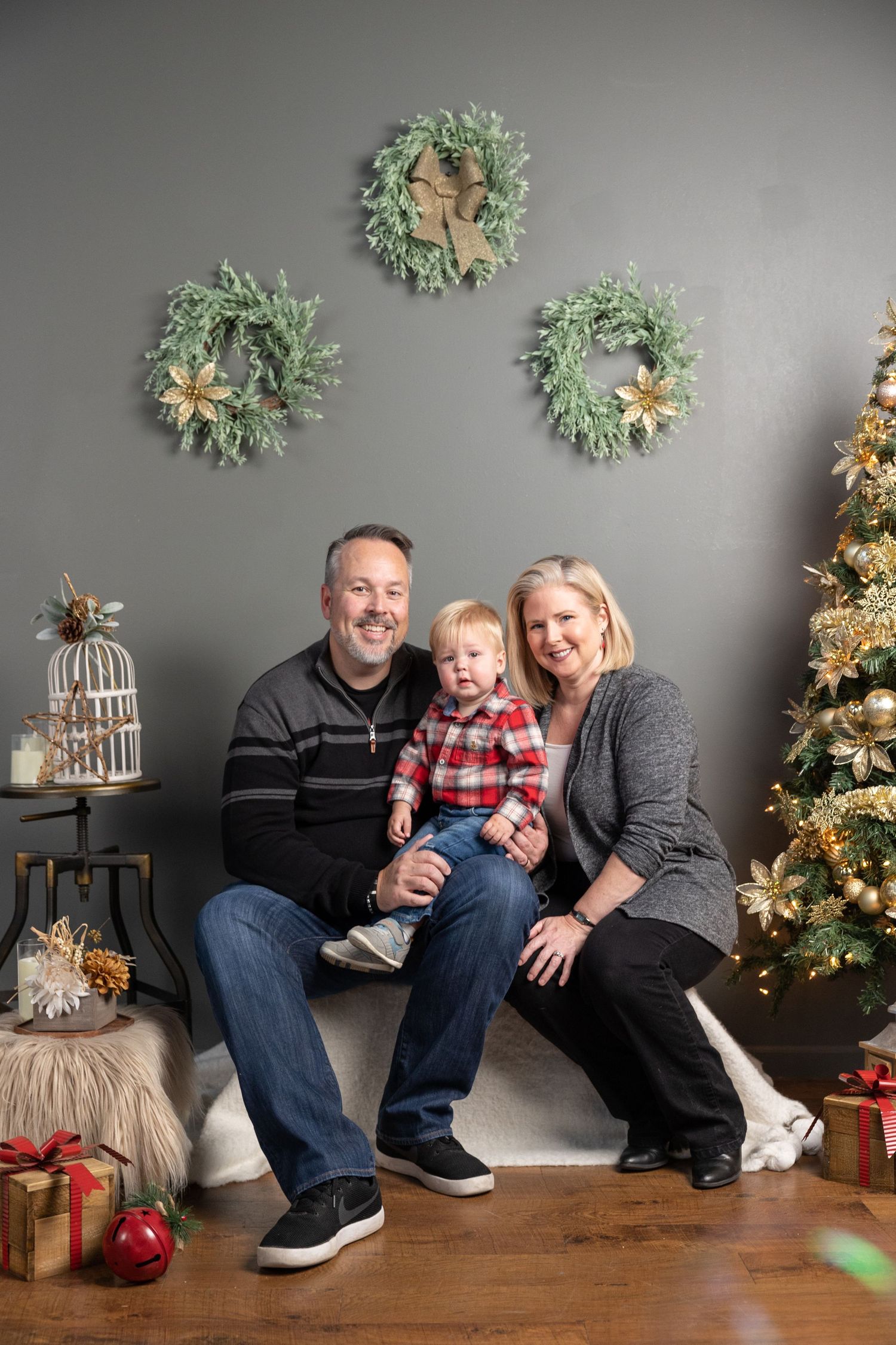 Family sits together on gray bench with Christmas decorations and wreaths on wall during holiday photo session.