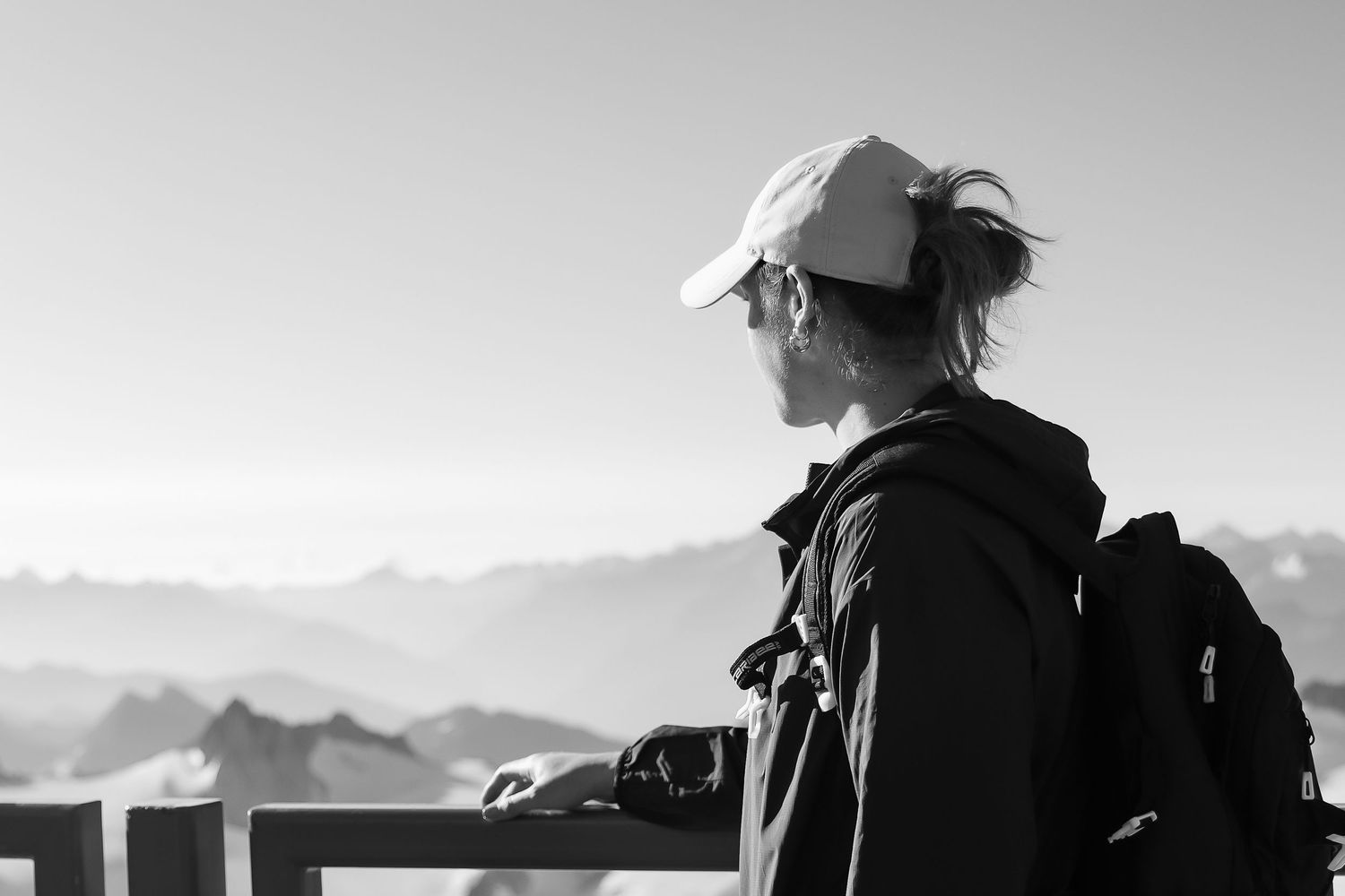 A hiker and their black and white dog sit atop a rocky mountain summit overlooking a vast valley landscape on a cloudy day.
