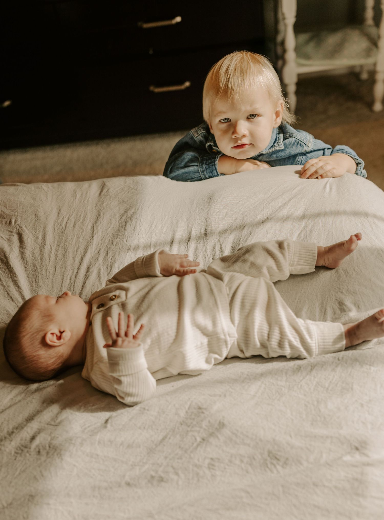 Baby in white outfit lying on cream bedding while sibling watches.