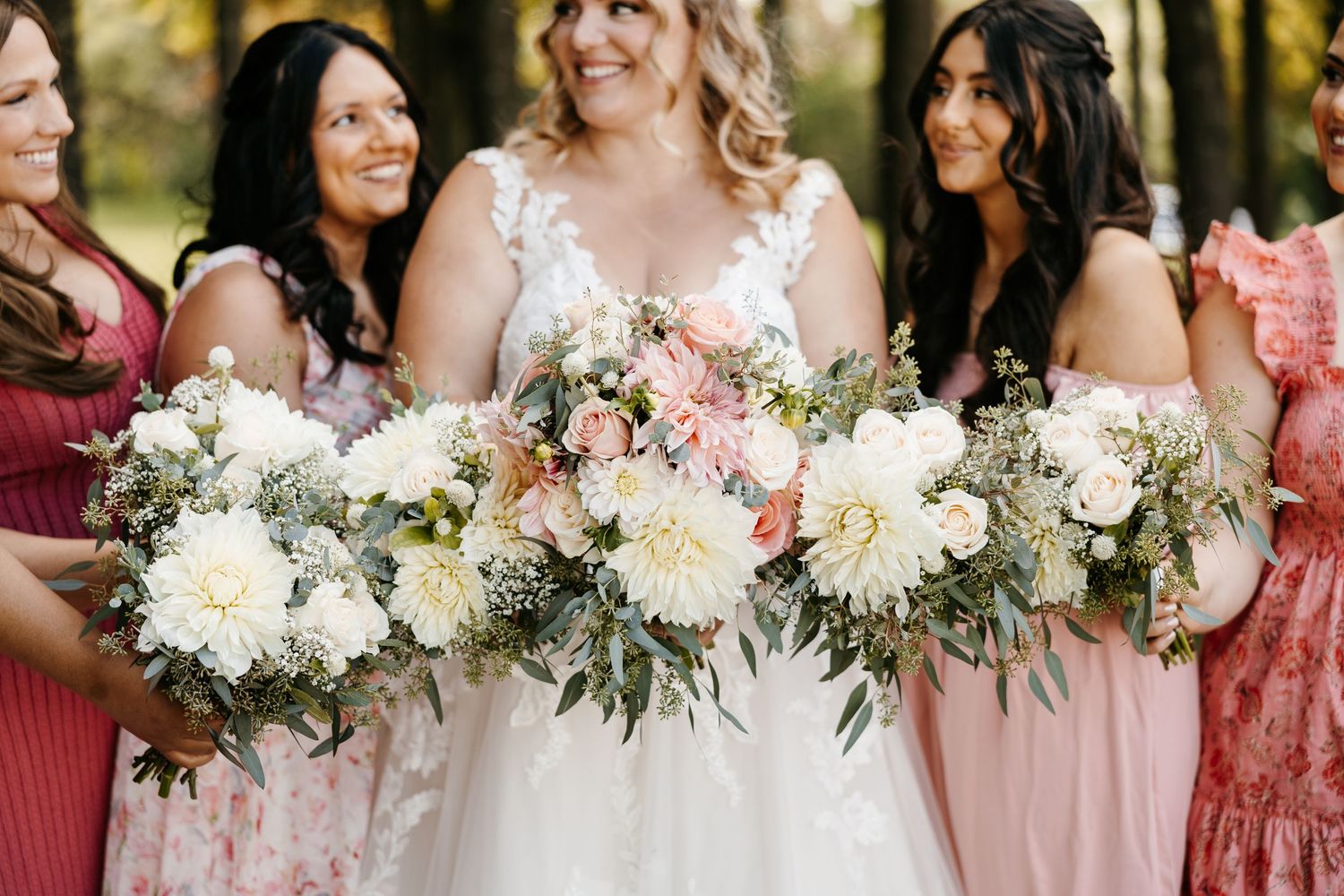 Bridesmaids in coral pink dresses hold elegant white and blush floral bouquets at an outdoor wedding ceremony.