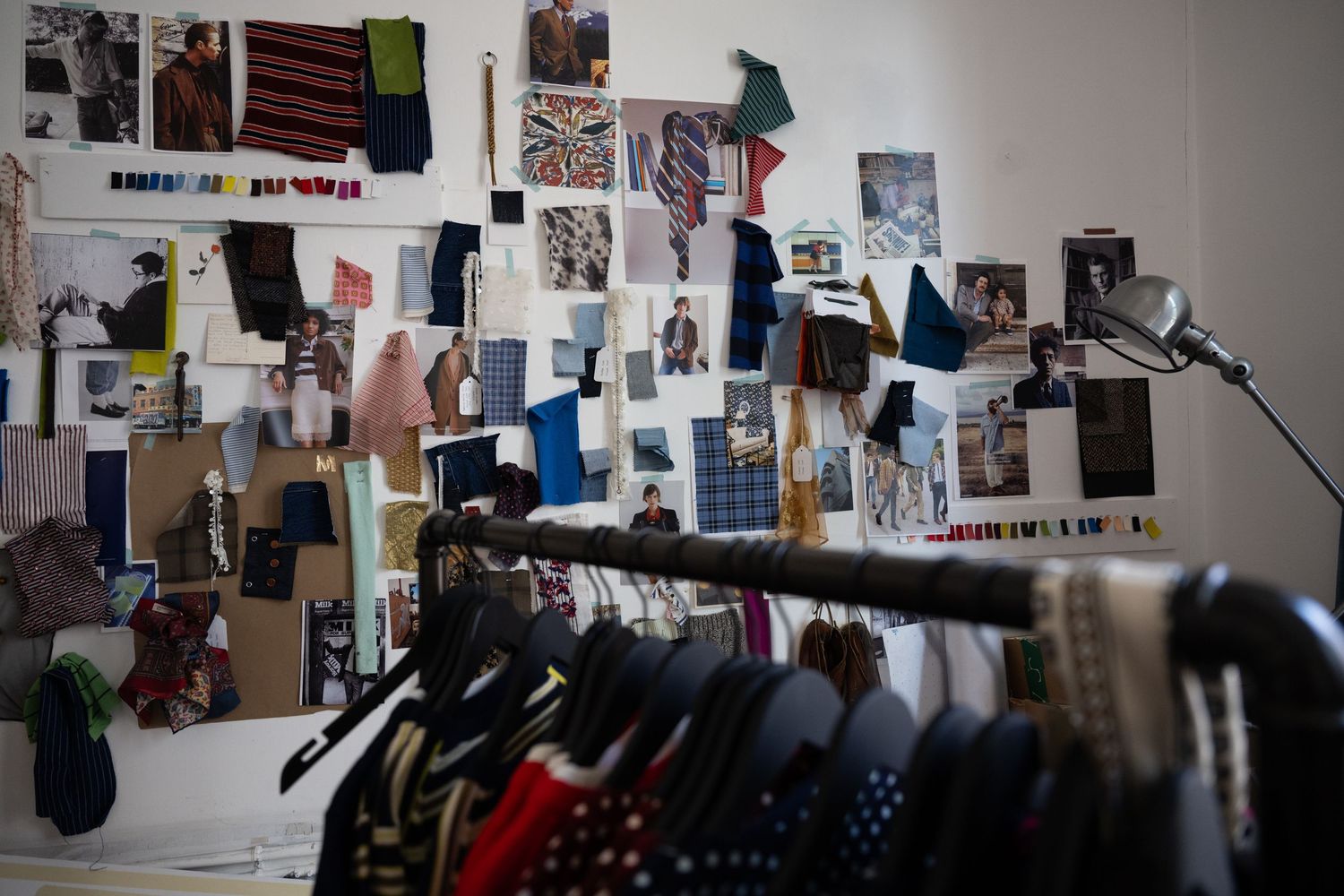 A cluttered workspace with photos and papers pinned to the wall and clothing items hanging on racks in a studio setting.