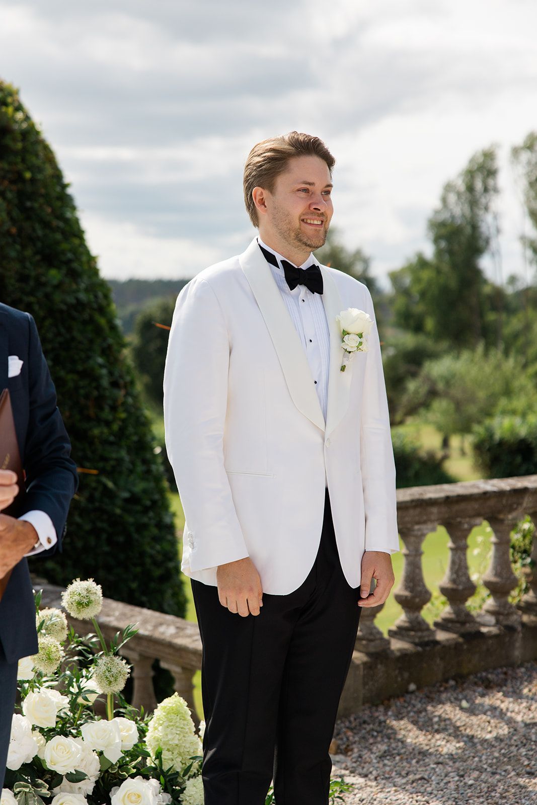 Wedding party members in black and white tuxedos stand on a stone terrace with elegant landscaping and greenery.