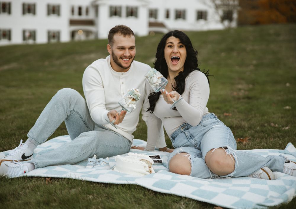 Excited couple sitting on blanket in park, laughing and holding gender reveal confetti announcement.