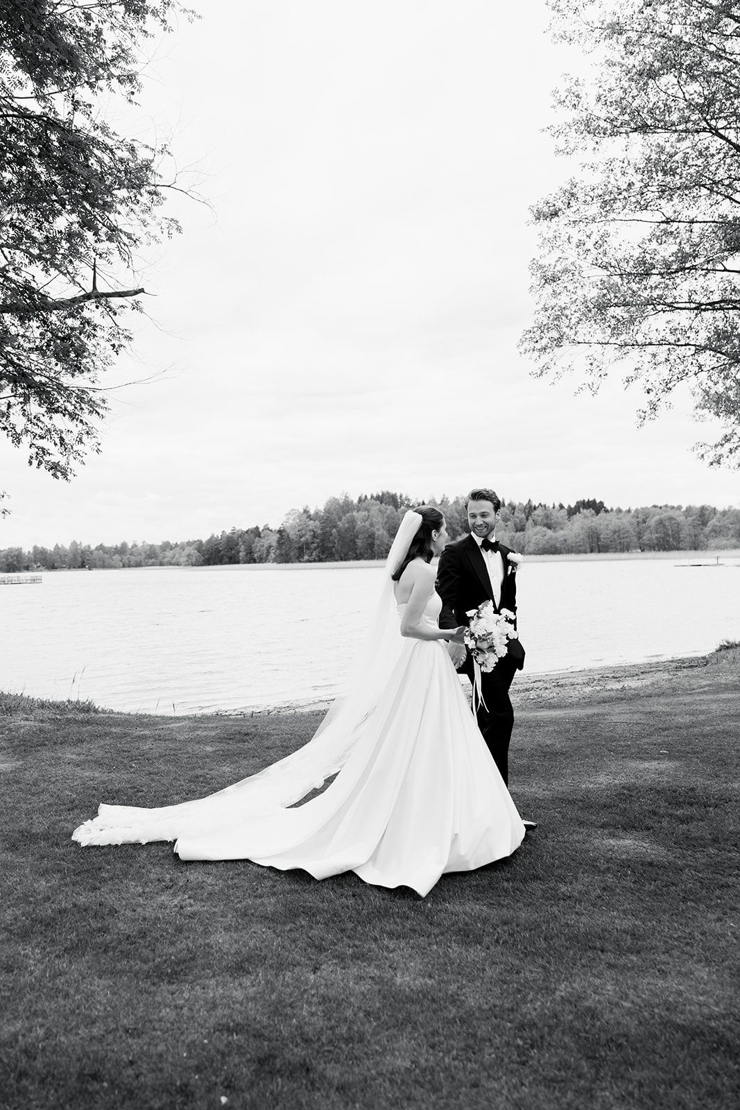 A couple in wedding attire dances together on a grassy hillside overlooking a lake in black and white photography.