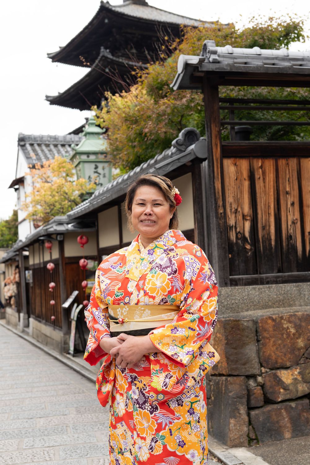 Woman in vibrant orange floral kimono poses on traditional Kyoto street with pagoda in background.