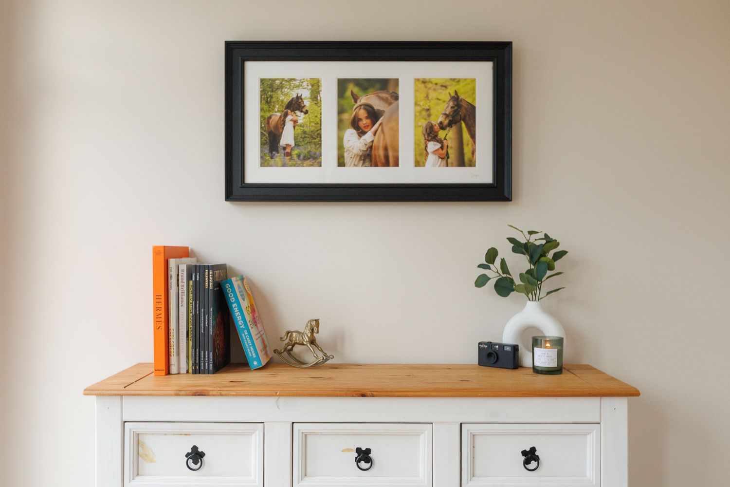 White painted wooden sideboard with dark hardware and wood top displays framed photos and flowers in natural lighting.