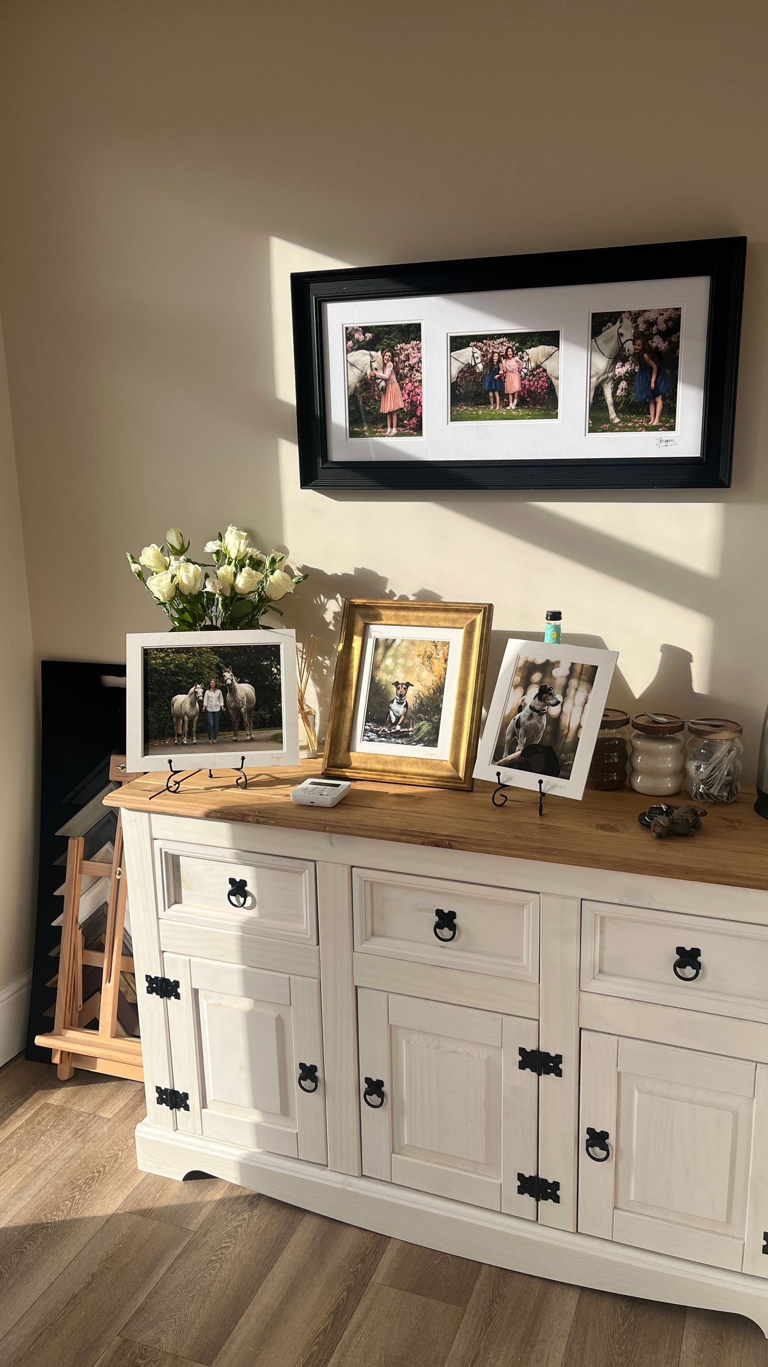 White painted wooden sideboard with dark hardware and wood top displays framed photos and flowers in natural lighting.