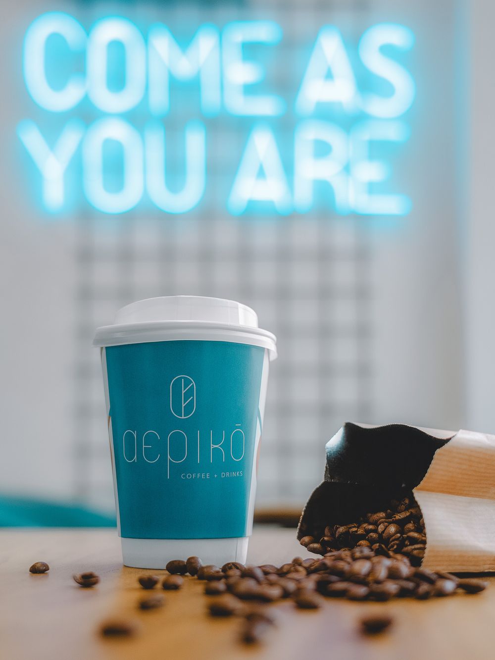 Coffee cup placed on a wooden surface with dessert, photographed below a neon sign reading “Come As You Are”