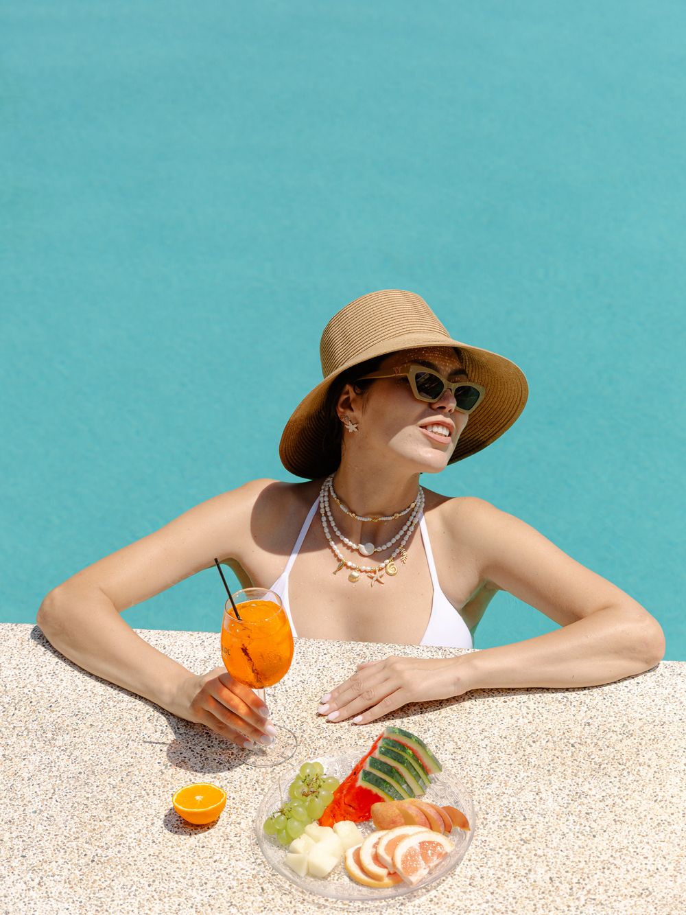 Woman partially submerged in a turquoise pool, resting her hands on the pool edge, wearing a sun hat and sunglasses under summer light.
