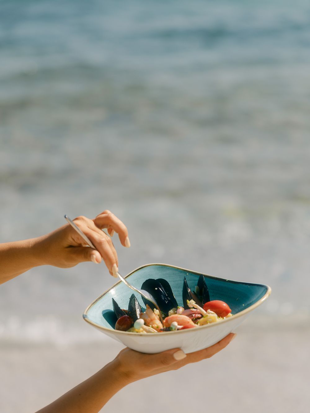 Hands holding a seafood bowl by the seaside with blue water in the background.