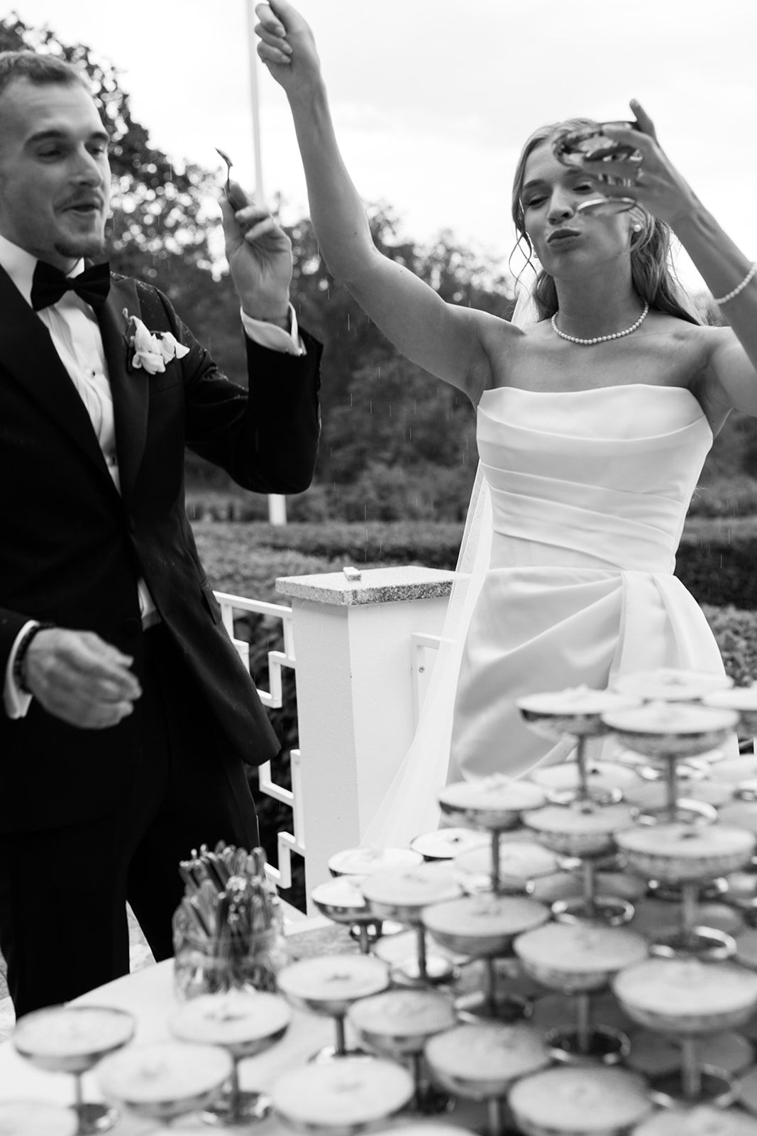 A black and white sequence of wedding guests celebrating while pouring champagne into a tower of glasses at a reception.