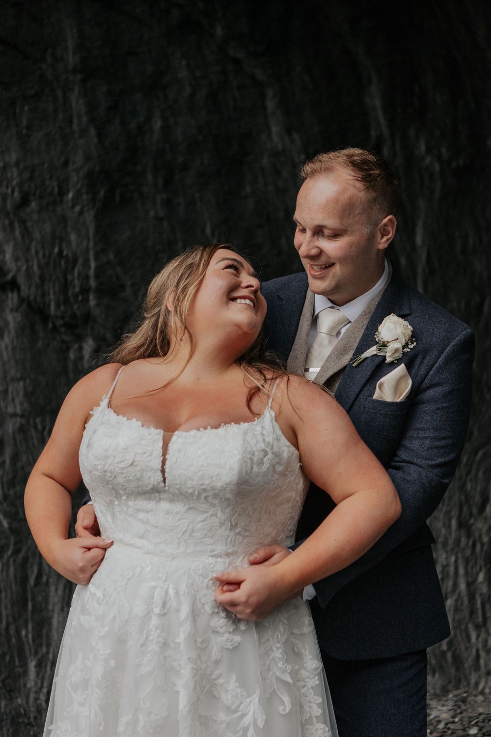 Bride and groom share a romantic moment embracing and smiling at each other against a dark rocky backdrop.