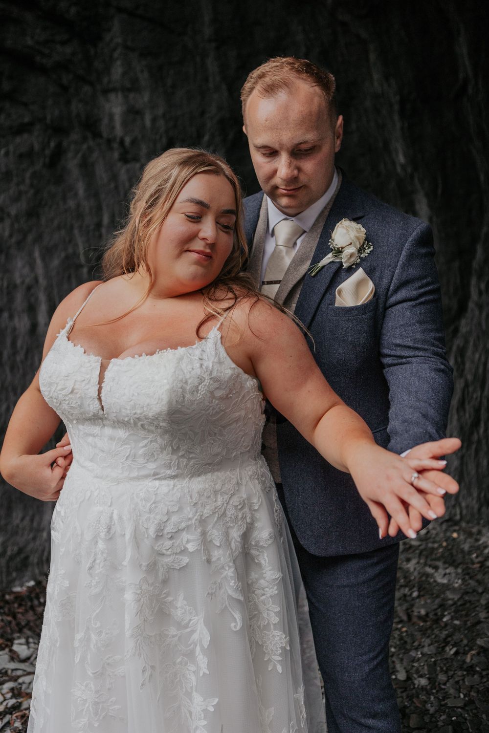 Bride and groom hold hands tenderly in an intimate pose against a dramatic dark stone background.