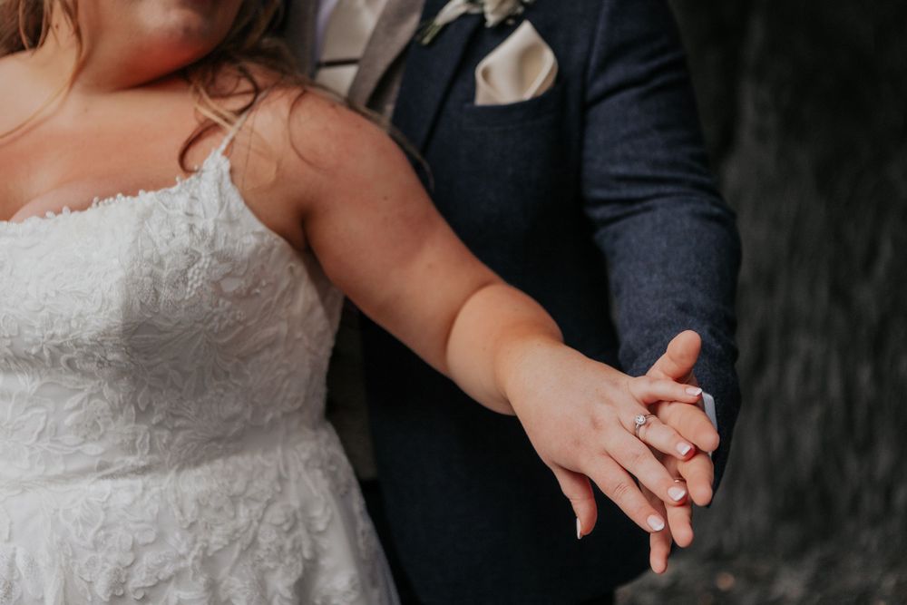 Close-up of bride and groom holding hands showing the wedding ring against a dark rocky background.