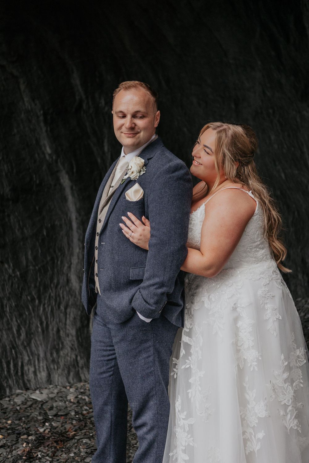 Bride leans lovingly against her groom as they pose together against a dramatic dark rocky backdrop.