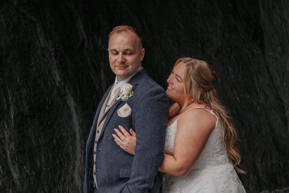 Bride in white lace gown embraces groom in grey suit with boutonniere against dark stone wall.