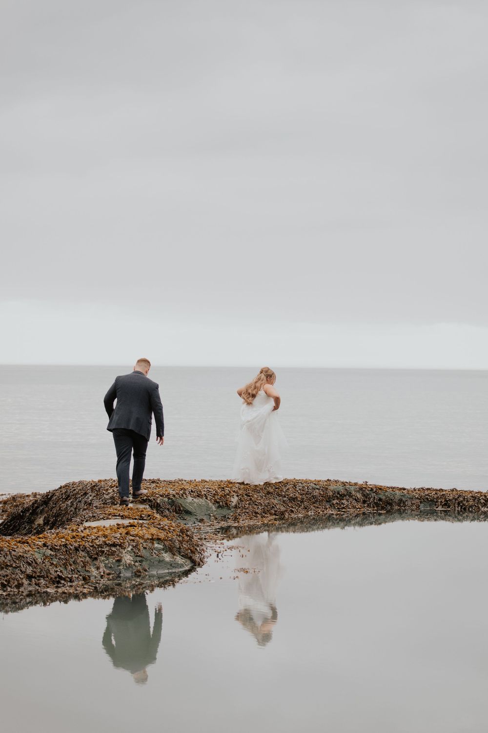 Bride and groom walk on rocky coastal shoreline with calm ocean reflection on overcast day.