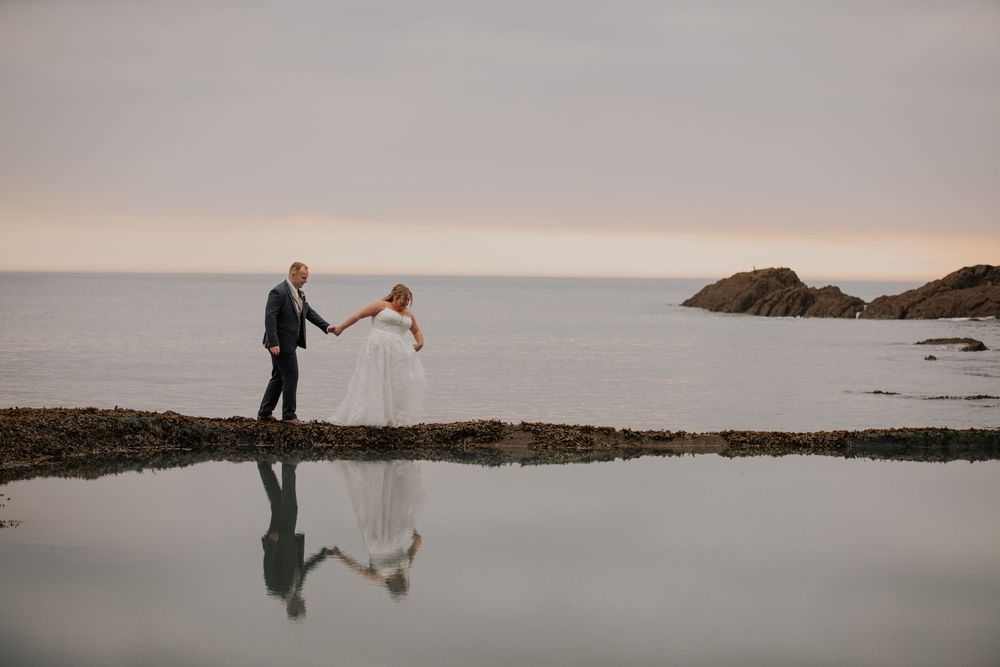 Bride and groom hold hands walking along rocky coast at sunset with mirror-like water reflection.