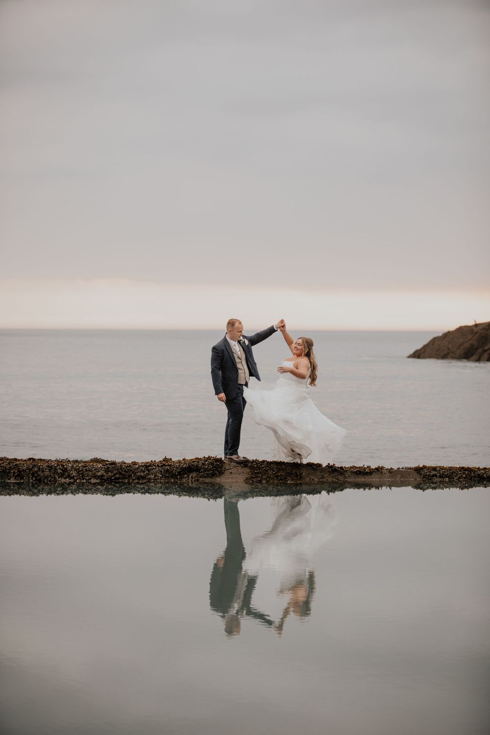 Groom twirls bride in flowing white gown on coastal rocks with stunning ocean reflection below.
