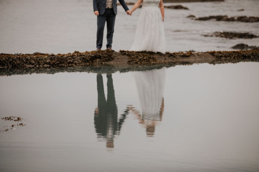Artistic reflection shot of bride and groom holding hands mirrored in calm coastal tidal pool.