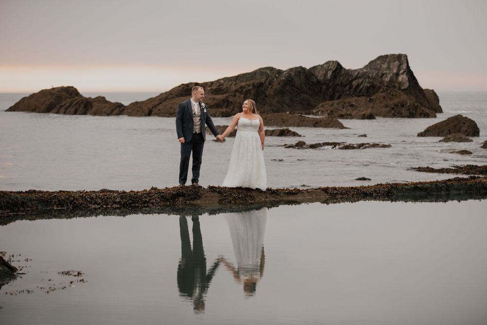 Bride and groom hold hands on rocky beach at dusk with dramatic rock formations and reflection.