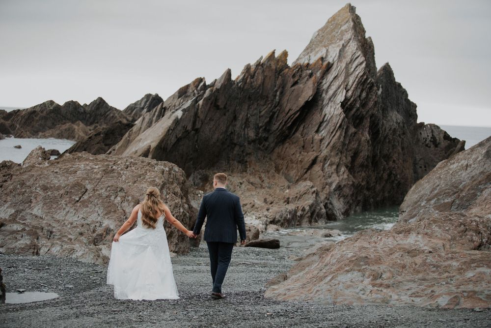 Bride and groom walk hand in hand toward dramatic pointed rock formation on rocky beach.