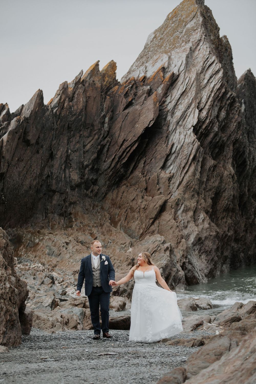 Bride and groom walk hand in hand before dramatic jagged rock formation on pebble beach.
