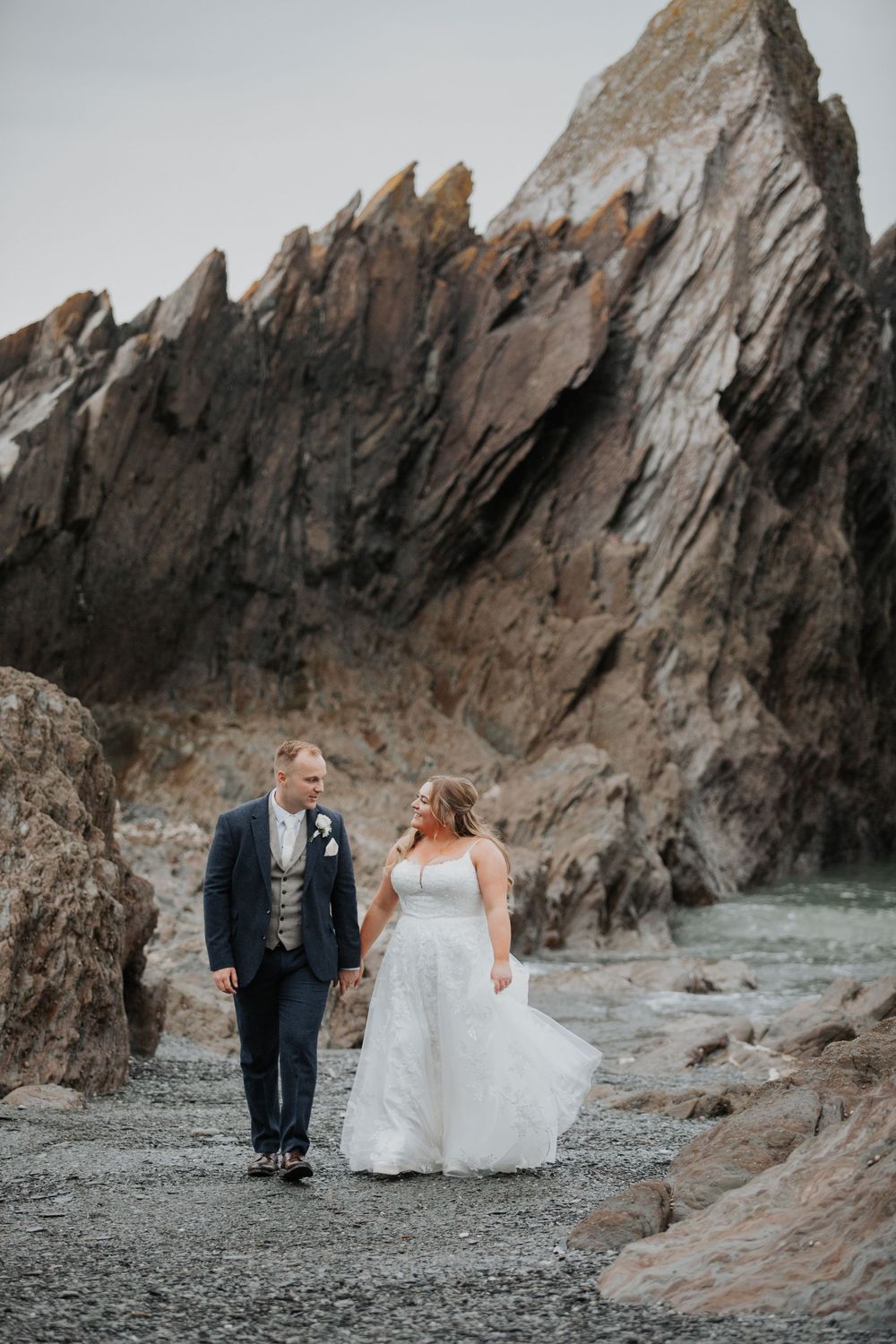 Bride and groom walk hand in hand on a rocky beach with dramatic cliffs behind them.