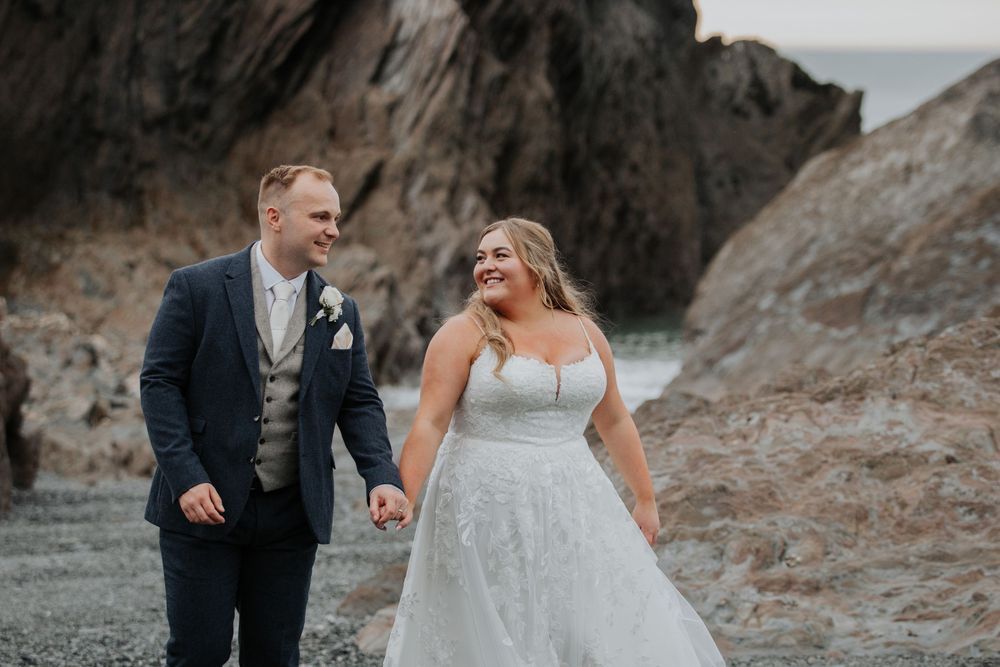 Smiling bride in lace gown and groom in navy suit hold hands on rocky coastal beach.