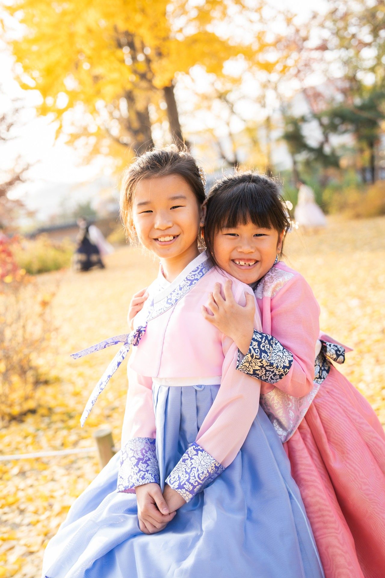 Two children in traditional Korean hanbok dresses play together among yellow autumn leaves in a park.