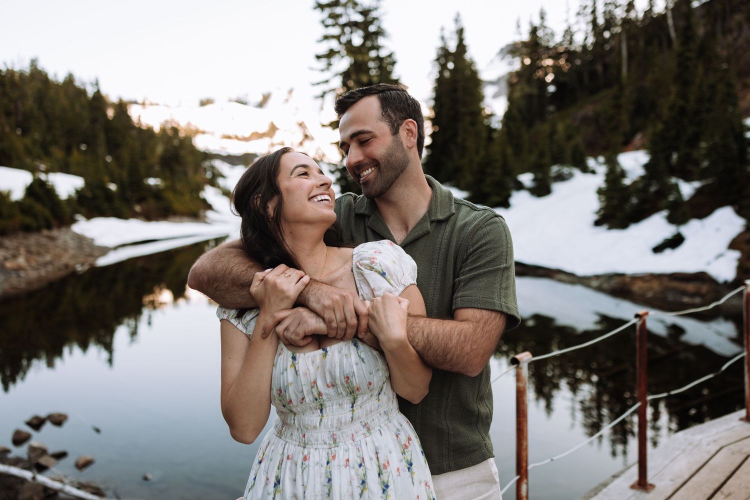 A couple shares romantic moments in a snowy mountain setting with evergreen trees and a reflective lake in the background.