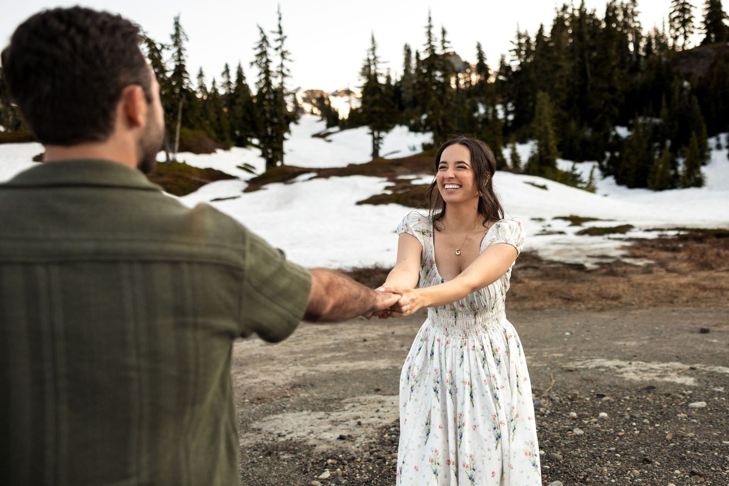 A couple dances together in a snowy mountain landscape at sunset with evergreen trees in the background.