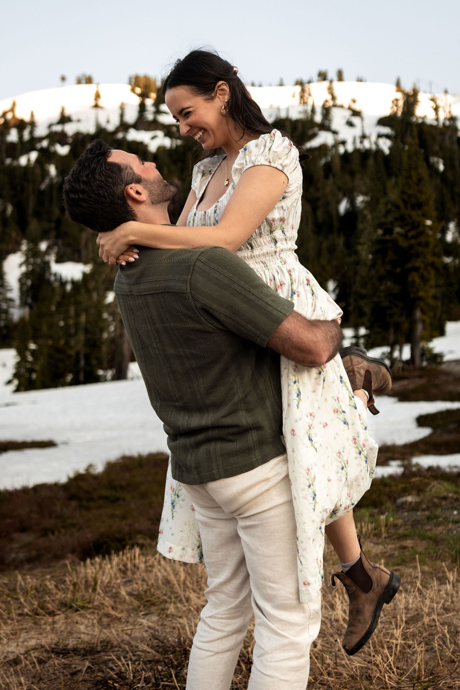 A playful couple embraces and laughs together against a scenic mountain landscape with patches of snow.