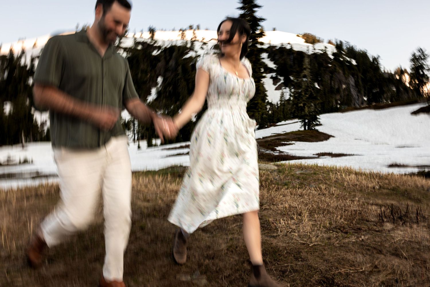 A couple in light colored outfits dances playfully together on a grassy hillside during sunset.