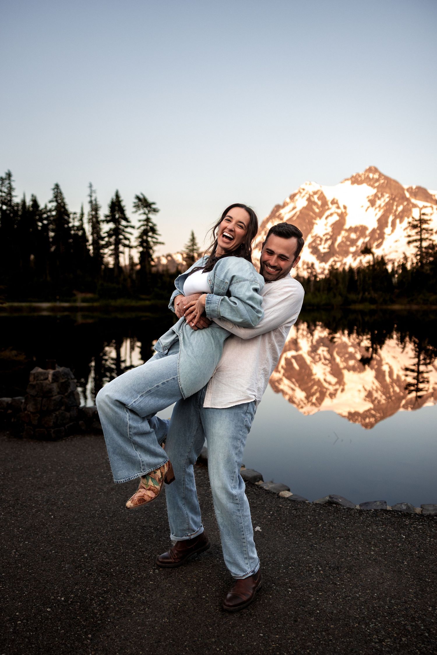 A playful couple dances and embraces on a rocky shore with snow-capped mountains glowing at sunset.
