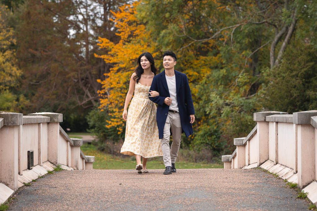 A couple walks along an elegant stone pathway surrounded by autumn trees in a romantic garden setting. Part of an engagement shoot.
