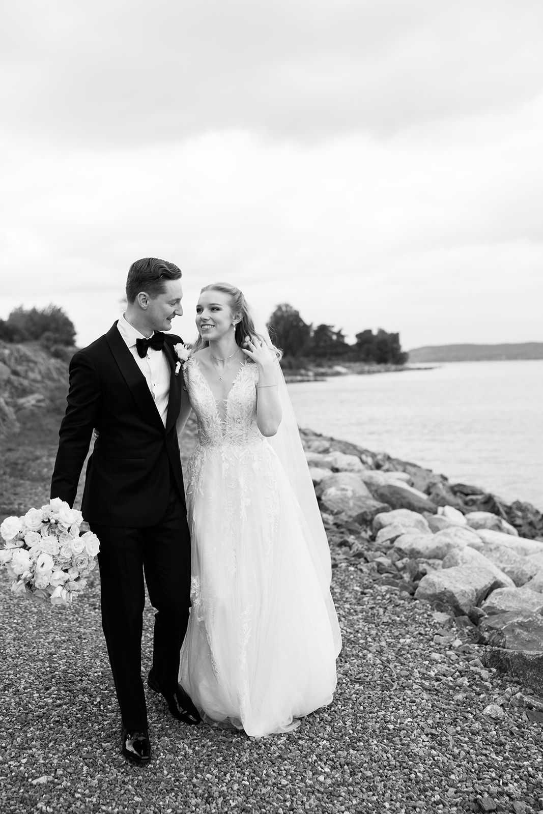 A couple in wedding attire walks along a grassy hill near a white church on an overcast day.