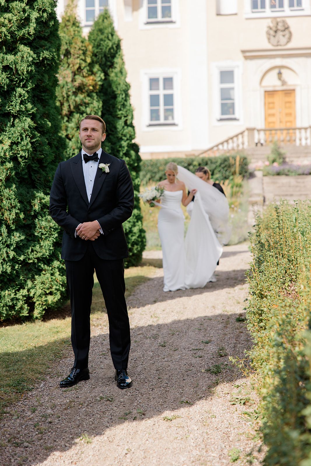 A groom in a classic black tuxedo stands on a garden path near evergreen trees and a stately building.